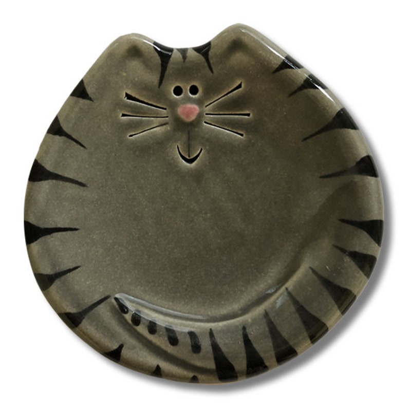 May include: A gray ceramic cat dish with black stripes and a pink nose. The dish is shaped like a cat with a smiling face.