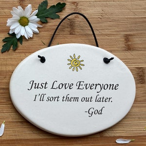 May include: White oval ceramic sign with a black string for hanging. The sign has a yellow sun with a swirl in the center and the text "Just Love Everyone I'll sort them out later. -God"