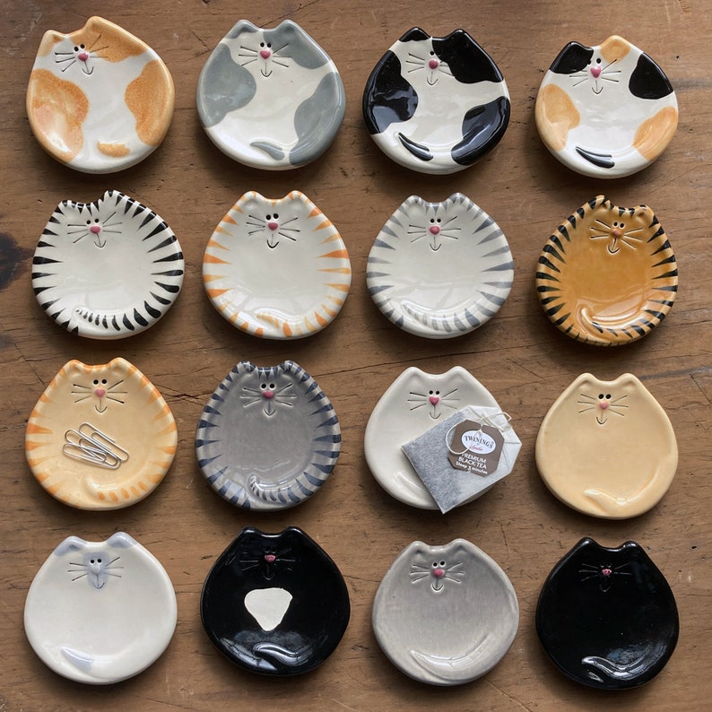 May include: Set of 16 ceramic cat teacup saucers in various colors and patterns. Each saucer features a cat face with a unique design.