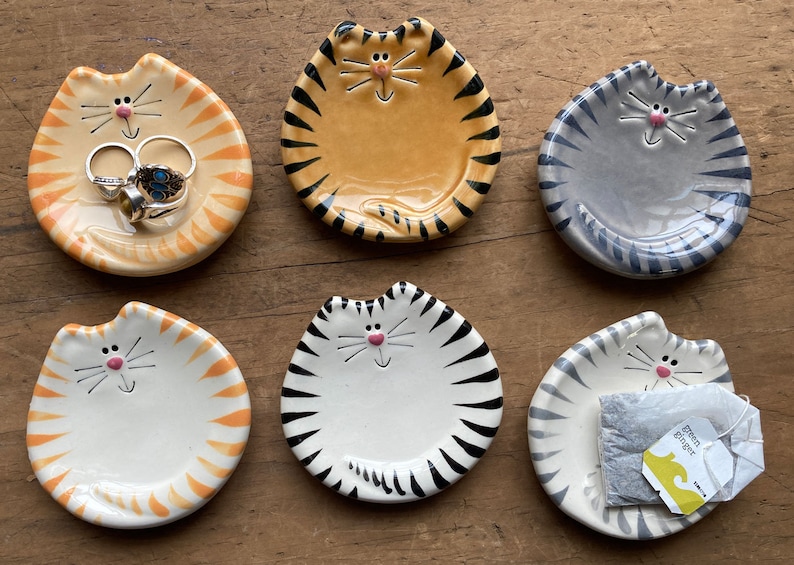 May include: Six ceramic cat-shaped trinket dishes with black stripes.  The dishes are in various colors, including orange, white, and gray.  One dish has a tea bag inside.