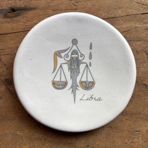 May include: White ceramic dish with a gray and gold illustration of the Libra zodiac sign. The sign is depicted as a scale with a stylized figure holding the scales. The word "Libra" is written below the image.