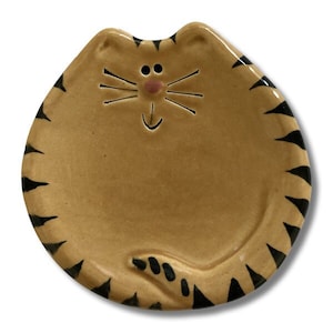 May include: A ceramic cat-shaped dish with black stripes and a pink nose. The dish is a light brown color and has a happy face.