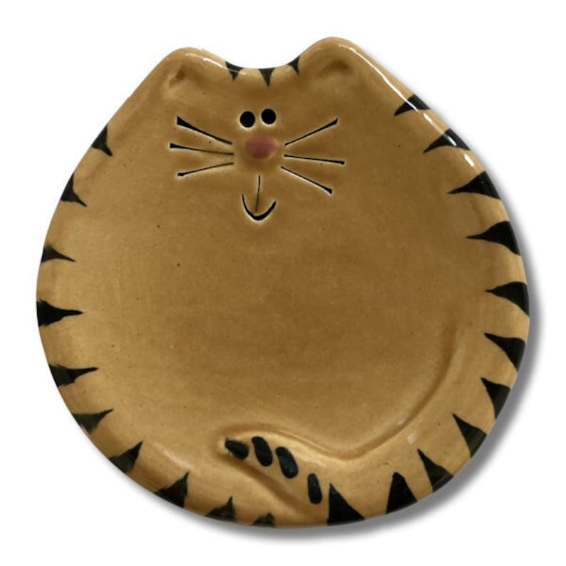May include: A ceramic cat-shaped dish with black stripes and a pink nose. The dish is a light brown color and has a happy face.