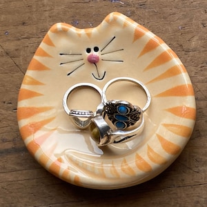 May include: A ceramic orange and white cat-shaped dish with a smiling face and whiskers. The dish is holding several rings with turquoise and gold stones.