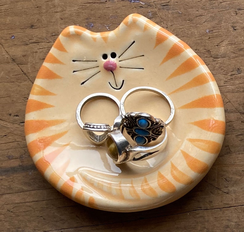 May include: A ceramic orange and white cat-shaped dish with a smiling face and whiskers. The dish is holding several rings with turquoise and gold stones.