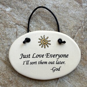 May include: White oval ceramic sign with a black string for hanging. The sign has a yellow sun with a spiral in the center and the text "Just Love Everyone I'll sort them out later. -God"
