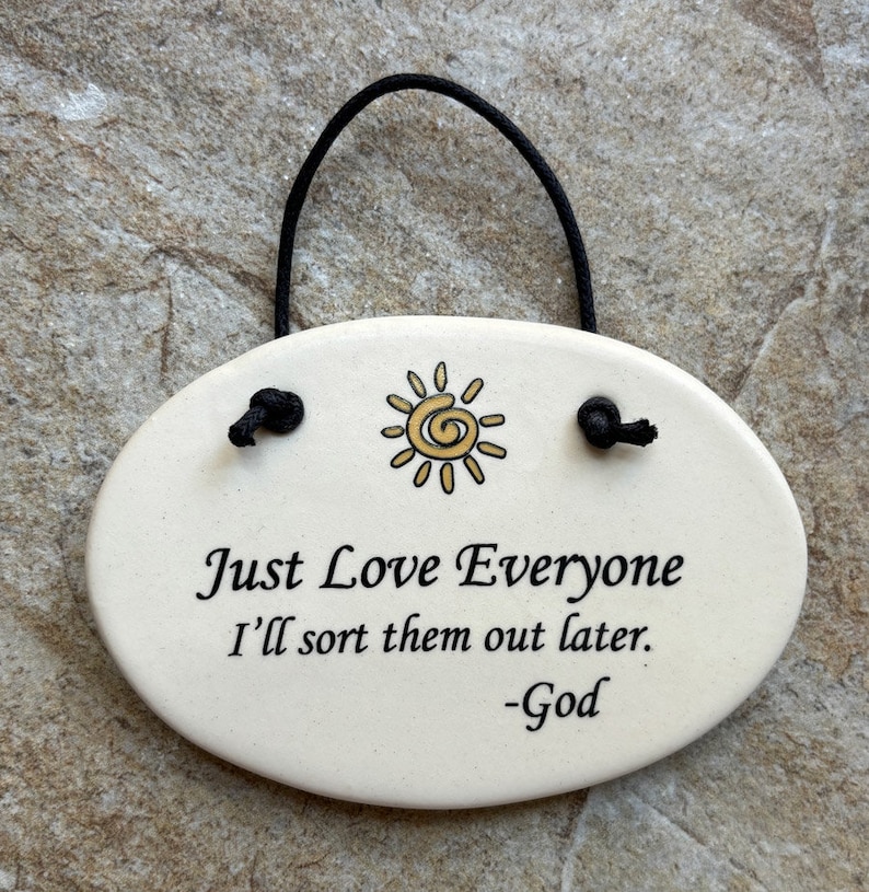 May include: White oval ceramic sign with a black string for hanging. The sign has a yellow sun with a spiral in the center and the text "Just Love Everyone I'll sort them out later. -God"