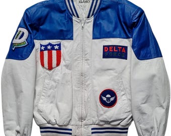 May include: White and blue bomber jacket with patches. The jacket has blue sleeves and a white body. Patches include a shield with stars and stripes, a Delta Force patch, and a patch with a parachute.