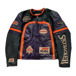 Puede incluir: Una chaqueta de moto de cuero negro y morado con rayas naranja y blancas. La chaqueta tiene varios parches, incluyendo "Goodyear", "Veedol", "IGOL" y "Skyjacker".