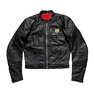 Może przedstawiać: Czarna sk&oacute;rzana kurtka motocyklowa z zamkiem błyskawicznym i złotą naszywką na lewej piersi. Kurtka ma pikowane detale na łokciach i ramionach.
