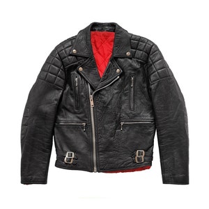 Op de afbeelding: Zwarte leren jas met een gewatteerd schouderontwerp en een rode binnenvoering. De jas heeft een zilveren ritssluiting, meerdere zakken en verstelbare taillebanden met gespen. Een klassieke jas in biker-stijl.