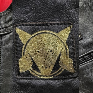 Może przedstawiać: Czarna sk&oacute;rzana kurtka z złotą naszywką przedstawiającą stylizowaną głowę zwierzęcia i okrąg z krzyżem w środku. Kurtka ma zamek błyskawiczny i kieszeń zapinaną na zamek błyskawiczny.