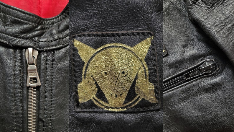 Może przedstawiać: Czarna sk&oacute;rzana kurtka z złotą naszywką przedstawiającą stylizowaną głowę zwierzęcia i okrąg z krzyżem w środku. Kurtka ma zamek błyskawiczny i kieszeń zapinaną na zamek błyskawiczny.