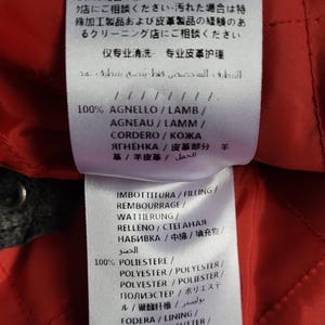 May include: A red fabric label with black text detailing the materials used in a garment. The label includes the words "100% Agnello/Lamb/Agneau/Lamm/Cordero/Кожа Ягненка/皮革部分羊 革/羊皮革/ الحمل" and "100% Poliestre/Polyester/Polyester/Polyester/Полиэстер/ポリエステル/聚酯纤维/بوليستر".