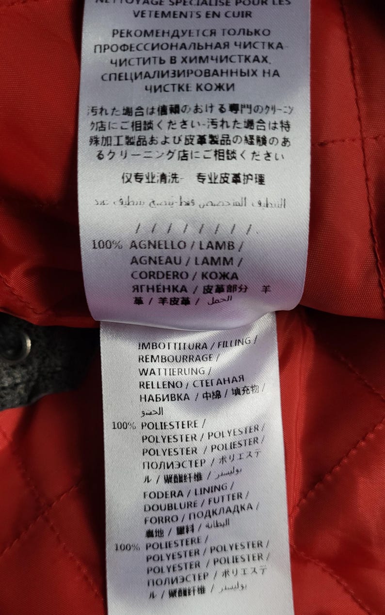 May include: A red fabric label with black text detailing the materials used in a garment. The label includes the words "100% Agnello/Lamb/Agneau/Lamm/Cordero/Кожа Ягненка/皮革部分羊 革/羊皮革/ الحمل" and "100% Poliestre/Polyester/Polyester/Polyester/Полиэстер/ポリエステル/聚酯纤维/بوليستر".