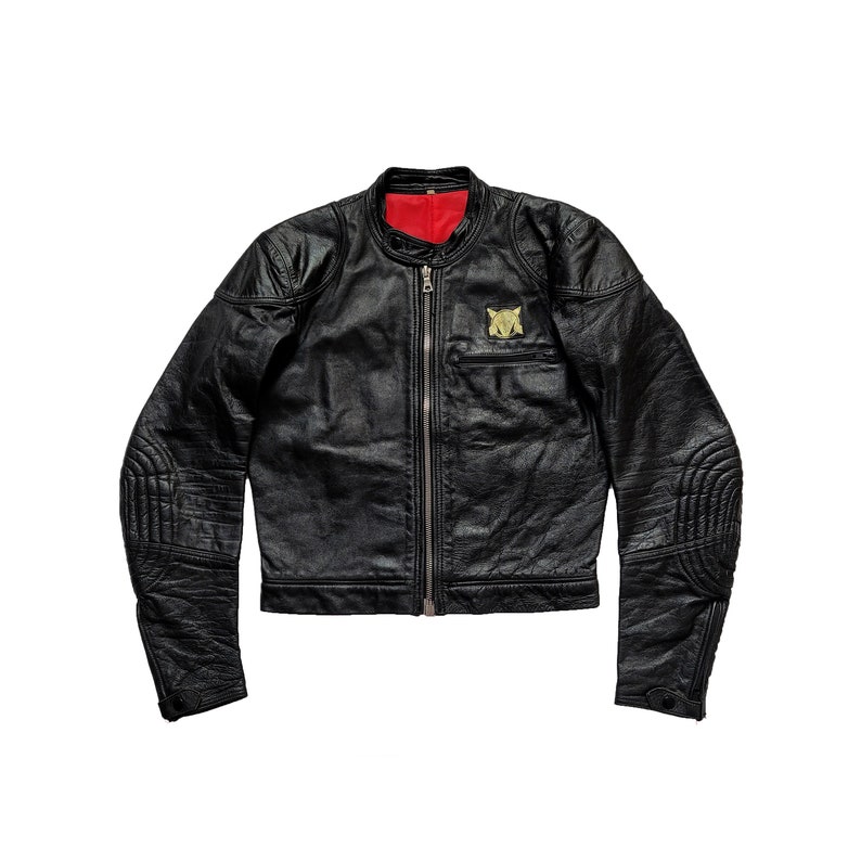 Może przedstawiać: Czarna sk&oacute;rzana kurtka motocyklowa z zamkiem błyskawicznym i złotą naszywką na lewej piersi. Kurtka ma pikowane detale na łokciach i ramionach.