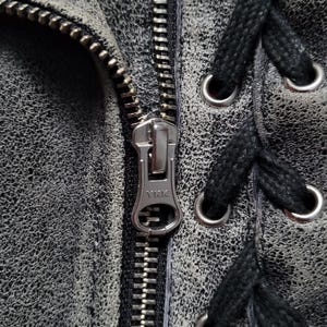May include: Close-up of a silver zipper on a black leather jacket with black laces and silver eyelets. The zipper has the text "YKK" on it.