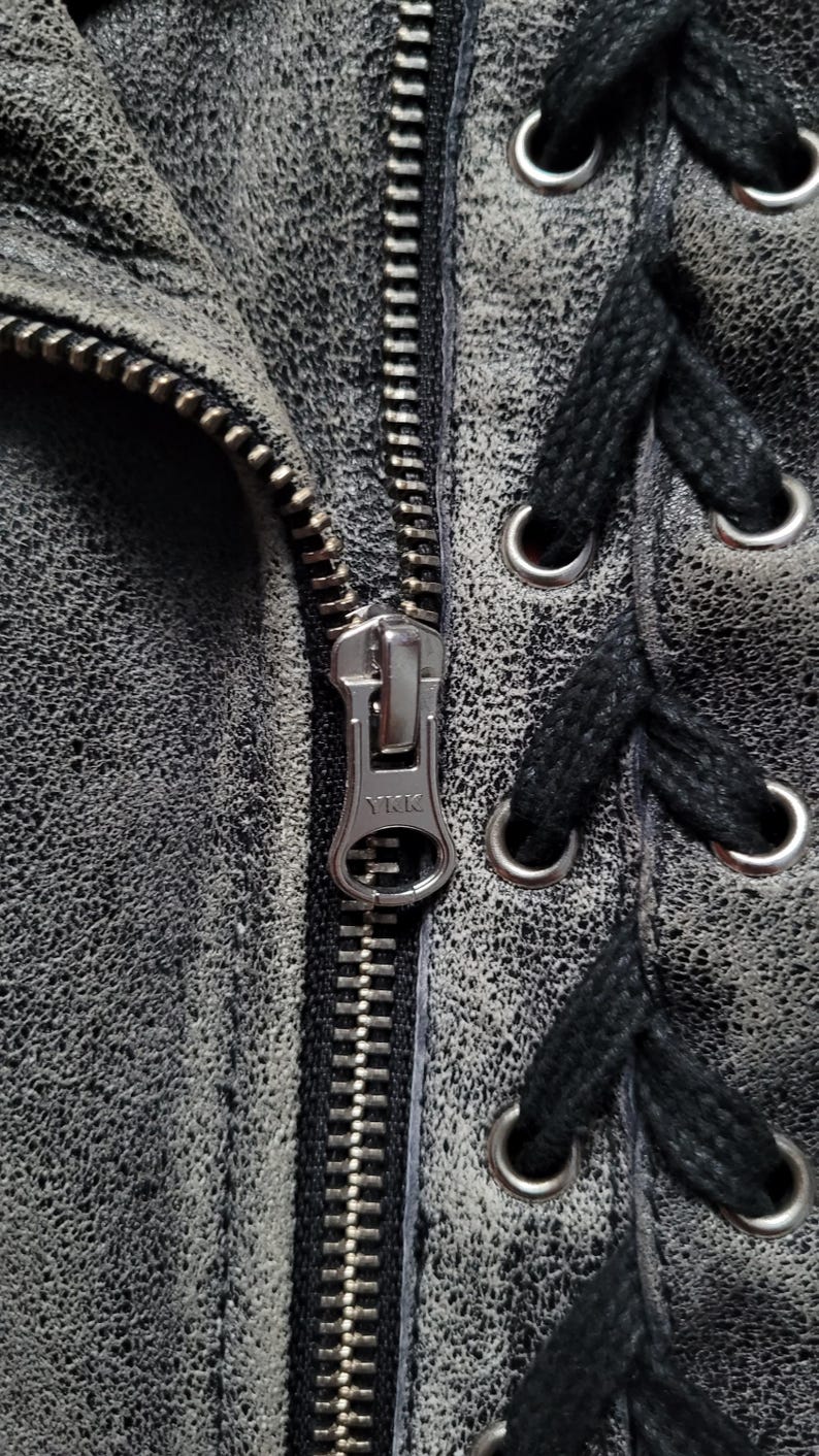 May include: Close-up of a silver zipper on a black leather jacket with black laces and silver eyelets. The zipper has the text "YKK" on it.