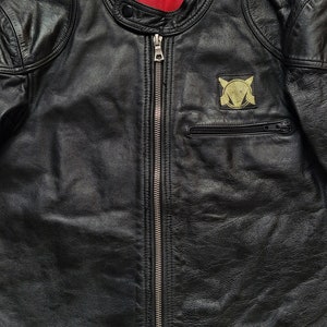 Może przedstawiać: Czarna sk&oacute;rzana kurtka motocyklowa z zamkiem błyskawicznym i złotą naszywką na lewej piersi. Naszywka przedstawia stylizowany wz&oacute;r z głową lisa i k&oacute;łkiem.