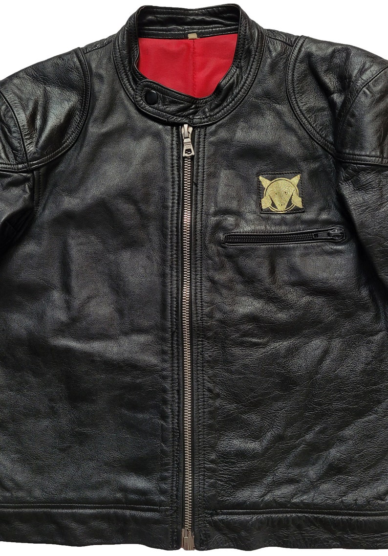 Może przedstawiać: Czarna sk&oacute;rzana kurtka motocyklowa z zamkiem błyskawicznym i złotą naszywką na lewej piersi. Naszywka przedstawia stylizowany wz&oacute;r z głową lisa i k&oacute;łkiem.