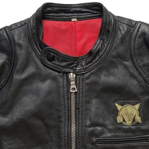 Może przedstawiać: Czarna sk&oacute;rzana kurtka z czerwoną podszewką i złotą naszywką po lewej stronie. Naszywka przedstawia stylizowany wz&oacute;r z k&oacute;łkiem i dwoma tr&oacute;jkątami.