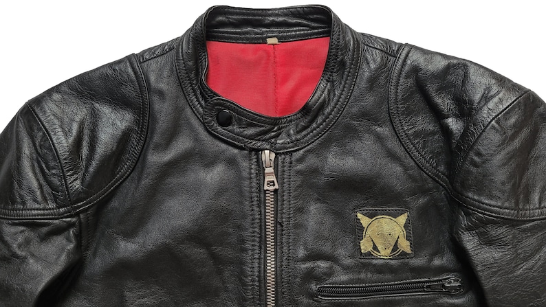 Może przedstawiać: Czarna sk&oacute;rzana kurtka z czerwoną podszewką i złotą naszywką po lewej stronie. Naszywka przedstawia stylizowany wz&oacute;r z k&oacute;łkiem i dwoma tr&oacute;jkątami.