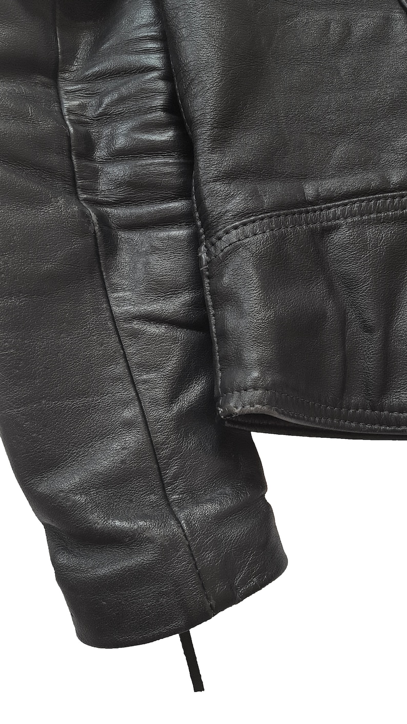 May include: Close-up of a black leather jacket sleeve with a seam detail. The sleeve is folded over and the fabric is textured.