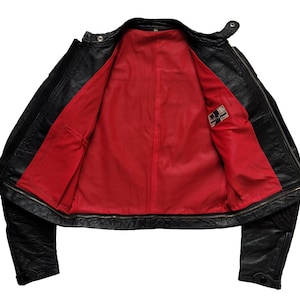 Może przedstawiać: Czarna sk&oacute;rzana kurtka z czerwoną podszewką i małą metką wewnątrz. Kurtka ma zamek błyskawiczny i dwie kieszenie z przodu.