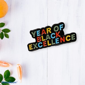 May include: Black enamel pin with the words "Year of Black Excellence" in rainbow colours. The pin is on a white wooden surface.