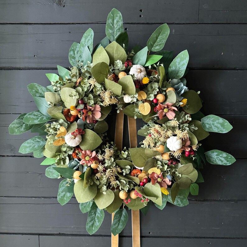 May include: A green, brown, and white fall wreath with a brown ribbon. The wreath is made of artificial leaves, berries, and flowers. The wreath is decorated with small white pumpkins and brown berries.
