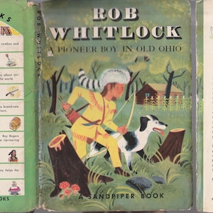 May include: A vintage hardcover book titled Rob Whitlock: A Pioneer Boy in Old Ohio. The green dust jacket features an illustration of a child in a coonskin cap with a bow and arrow and a dog, alongside a list of other Sandpiper Books titles.
