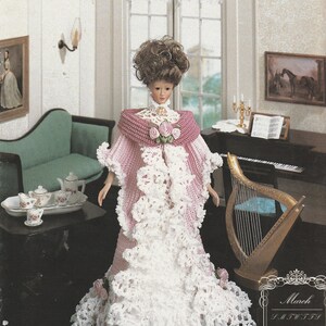 May include: A doll wearing a floor-length gown featuring a pink crocheted bodice and a white crocheted lace skirt with ruffled trim. The outfit includes a pink crocheted shawl adorned with three pink fabric roses at the center neckline.