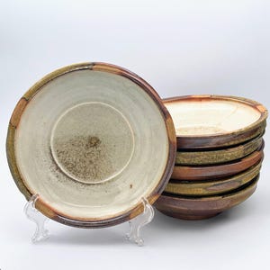May include: A stack of handmade ceramic plates with a rustic aesthetic. The plates feature a light beige interior, a brown rim, and a textured, earthy exterior. One plate is displayed on a clear stand, showcasing the design.