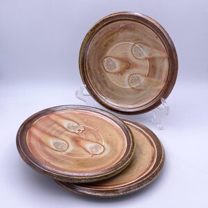 May include: A set of three handmade ceramic plates. The plates have a light brown center with reddish-brown streaks and a dark brown rim. One plate is elevated on a clear stand, while the other two are stacked. The plates have a rustic, earthy aesthetic.