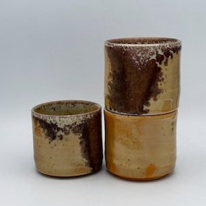 May include: Three cylindrical ceramic cups with a smooth, light tan base glaze. The upper sections feature a mottled, dark brown drip pattern that cascades down the sides. The cups have a simple, handleless design with a consistent, straight-walled shape.
