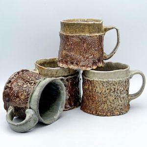 May include: Four handmade ceramic mugs with textured, bark-like exteriors in shades of brown and tan. Each mug has a unique shape and handle, with a light-colored rim and interior. The mugs are stacked and arranged on a white background.