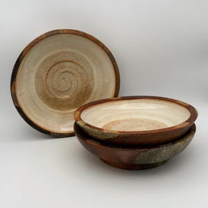 May include: Three ceramic bowls with a cream-colored interior and a dark, textured brown exterior. The bowls feature a visible spiral pattern at the base of the interior and a thick, rounded rim. The finish appears matte and earthy in tone.