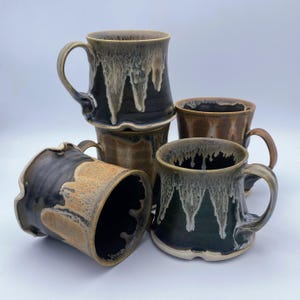 May include: A collection of handmade ceramic mugs in various shades of brown and black. Each mug features a unique dripped glaze design in white and tan, with a curved handle. The mugs are stacked and arranged to showcase their individual shapes and textures.