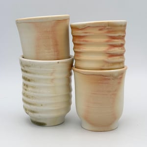 May include: Four handmade ceramic cups in a stack. The cups are off-white with reddish-brown and olive green accents. Each cup has a unique textured design, with some featuring horizontal ridges and others a smooth, curved shape. The cups are stacked on a white background.