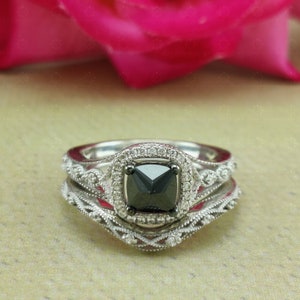 May include: A three-piece engagement ring set with a black square-shaped diamond surrounded by smaller diamonds in a halo setting. The band is a delicate, intricate design with smaller diamonds.