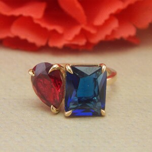 Könnte beinhalten: Ein goldener Ring mit einem roten herzförmigen Stein und einem blauen quadratischen Stein.