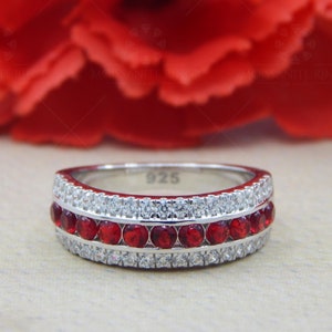 Pode incluir: Um anel de banda de prata com uma fileira de pedras preciosas vermelhas e duas fileiras de pedras preciosas transparentes. O anel é marcado com "925" no interior.