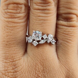 May include: A silver ring featuring a cluster of clear, sparkling gemstones. The ring has a central, round-cut stone surrounded by smaller, pear-shaped and round-cut stones, creating a floral or leaf-like design. The ring is set on a silver band.
