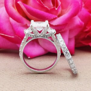 May include: A set of two silver engagement rings with a large square-shaped diamond in the centre of the engagement ring and smaller diamonds on the band. The rings are set against a pink rose background.