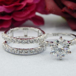 May include: A set of two silver rings with a large, round, clear diamond in the center of the top ring. The bottom ring is a simple band with small, clear diamonds.