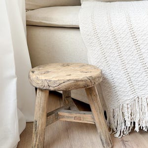 Peut inclure: Un petit tabouret en bois rustique avec un dessus rond et trois pieds. Le tabouret est en bois vieilli, mettant en valeur le grain et les fissures naturels. Une couverture crème à franges est drapée sur un canapé beige, créant une scène aux tons neutres et confortables.
