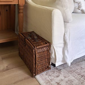 May include: A rectangular brown wicker picnic basket with dark brown leather straps and metal buckles, positioned on a light wood floor next to a white fabric sofa with matching cushions and a wooden hutch with a white ceramic pitcher.