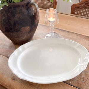 May include: A brown ceramic vase with white hydrangea flowers and green leaves. A white, oval serving platter with a scalloped edge is on a wooden table. A small glass candle holder with a lit candle is also on the table.