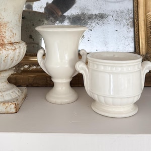 May include: A collection of three ceramic vessels including a weathered, rust-spotted urn containing a dark-bristled brush and a thin paintbrush, alongside two smooth, cream-coloured urns with side handles and pedestal bases.