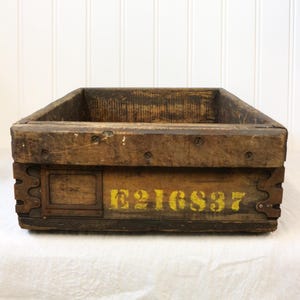 Peut inclure: Une caisse rectangulaire en bois avec une finition brune patinée, dotée de renforts d'angle en métal et du texte jaune au pochoir E216S37 sur le panneau avant. La boîte présente des veines de bois visibles, des taches sombres et des vis en métal.